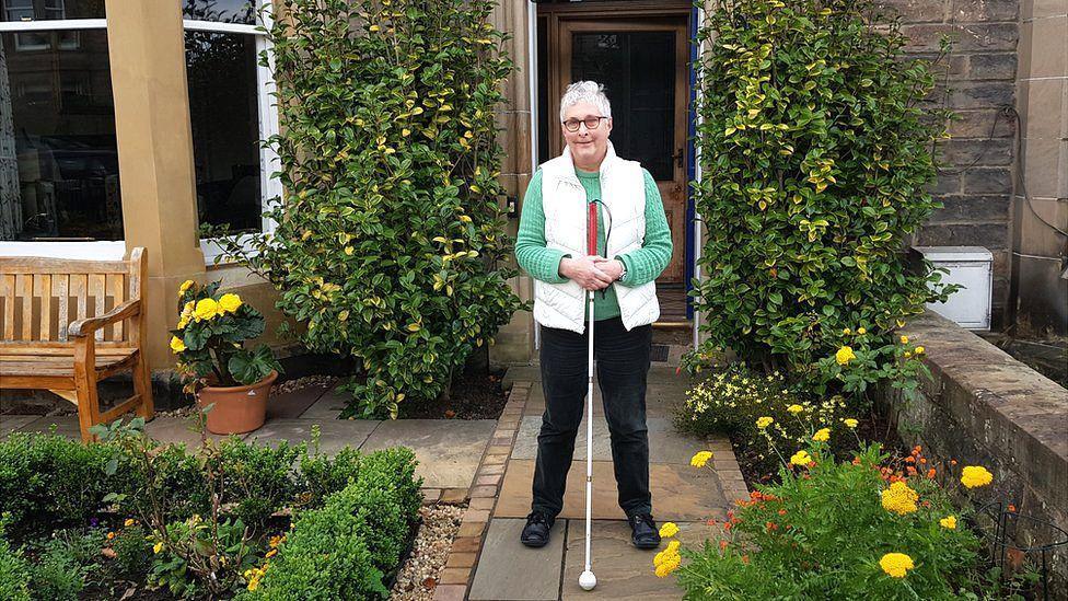 Lorna Pratt standing in a well-kept garden that has bushes and yellow flowers and a creeping plant that is growing up both sides of the front of the house she is outside. She is standing on a path that leads to the front door. She is holding her white cane and is wearing a white body warmer, black trousers and glasses.