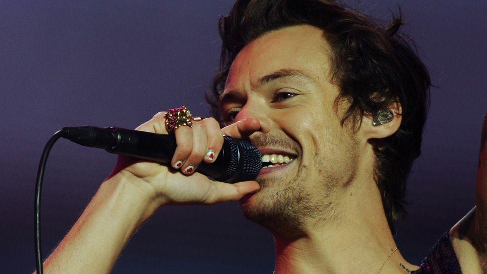 Harry Styles, with short dark hair, painted nails and a large gold ring, holds a microphone to his smiling mouth