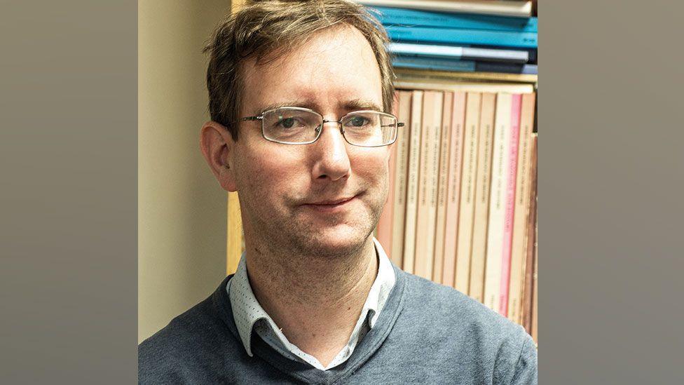 Edward Biddulph who has short light brown hair and is wearing metal-framed glasses and a blue v-necked jumper over a white shirt. Behind him is a row of reports.
