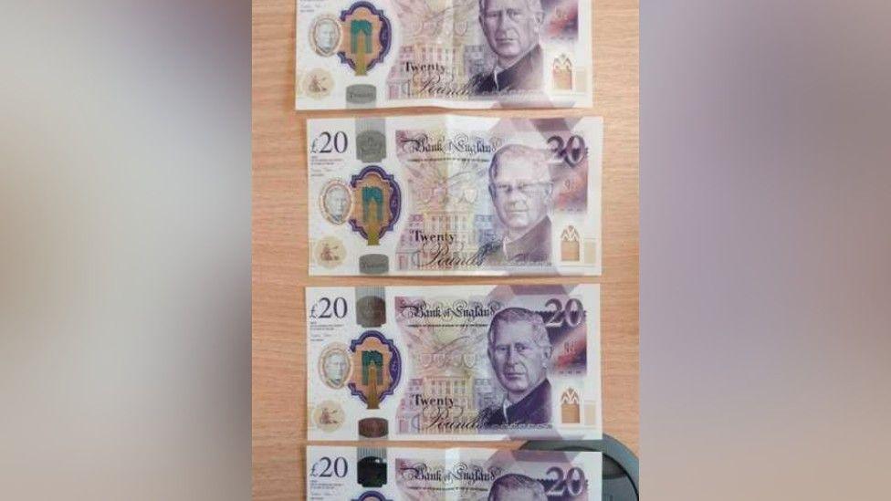 Four counterfeit £20 - bearing King Charles' image - laid out on a pine-coloured desk