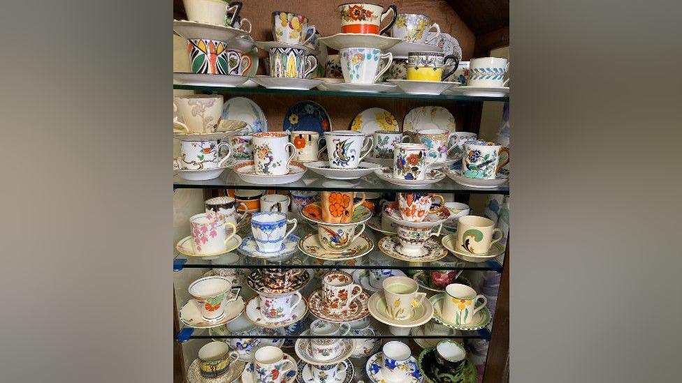 An open wood cabinet with glass shelves. There are dozens of brightly coloured ceramic coffee cups with matching saucers. Some are stacked on top of one another