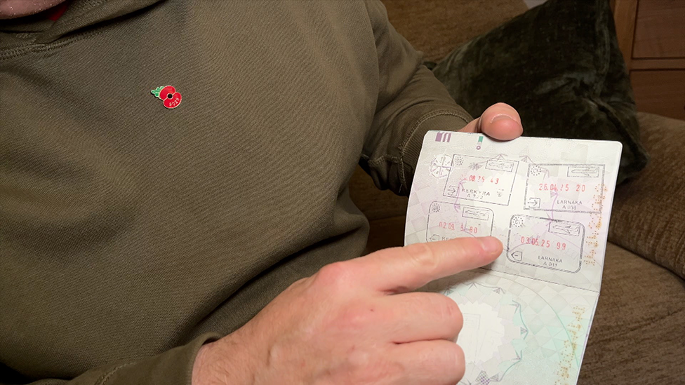 A man in an olive green jumper pointing to a page in a passport showing stamps for Lanarka on 26 April 2025 and 3 May 2025