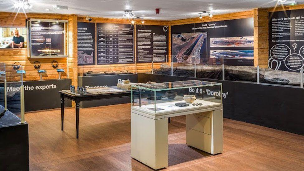 A view of displays at Flag Fen Archaeological Park. It shows a pine clad interior covered in black displays boards with white writing and photographs on the walls. Against the walls are black display cabinets with artefacts within. Against the wall on the right is a long preserved log boat. In the middle of the room are two more display cabinets with artefacts under glass and replica tools. 