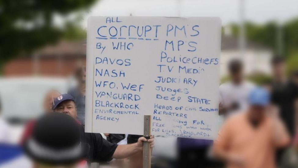 A man, wearing a blue England football hat, holds a white placard with blue writing. The writing says government ministers and MPs are corrupt, as are police chiefs, journalists, judges and the deep state, among a lot of other agencies.