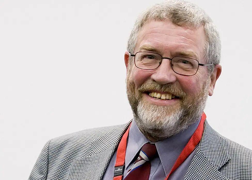 Brian Lomax in a suit and tie, smiling at the camera. He has a beard and short grey hair and is wearing glasses.