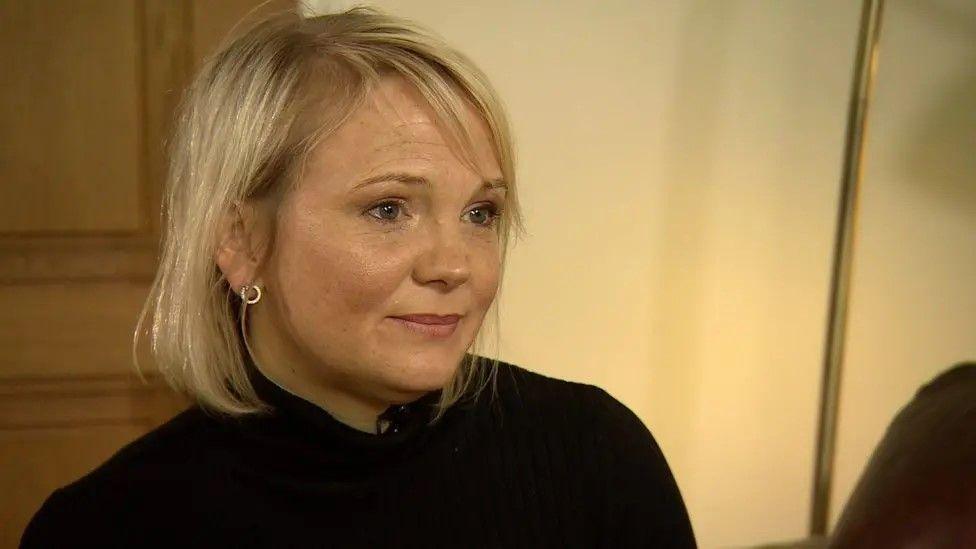 Elizabeth Hudson, a woman in her forties, with blonde hair, circular silver earrings, and a black turtle neck. It is a close-up shot of her head and shoulders, seemingly in her home or a living room.