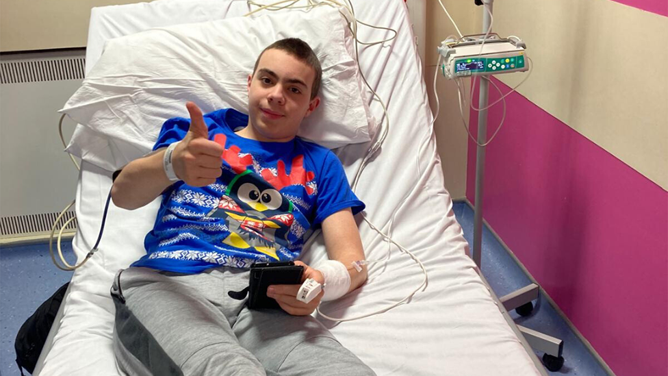 Sam lying in a hospital bed with various hospital kit attached to him. He is wearing a blue T-shirt and has his thumb up