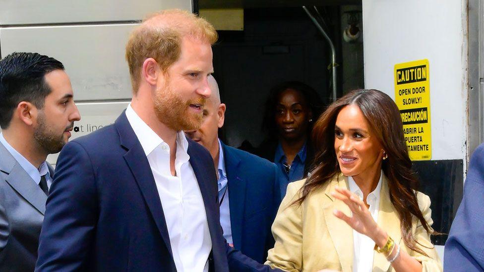 The Duke and Duchess of Suffolk walk out of a door while smiling. They are both wearing suits. Prince Harry is wearing a black blazer and trousers while Meghan is wearing a beige blazer and trousers. Both are wearing a white shirt. Prince Harry has ginger hair and blue eyes. Meghan has long brown hair and brown eyes.