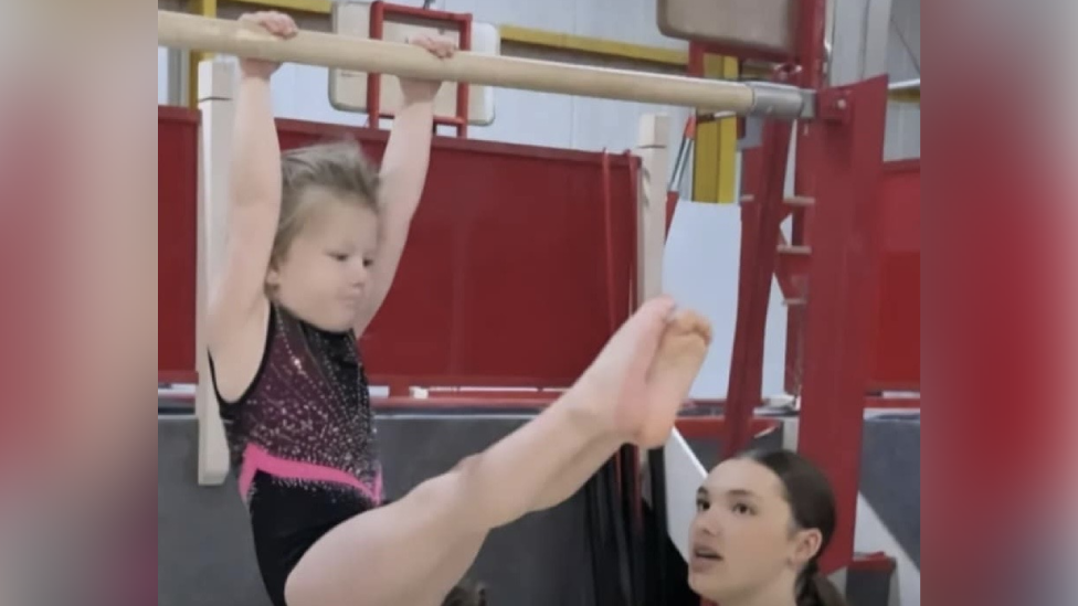 A young girl with tied-back blonde hair wearing a black and pink sparkly gymnastics leotard and swinging from a wooden bar with red gym apparatus behind her. Below her legs, you can see the head of a female instructor who has tied-back brown hair.