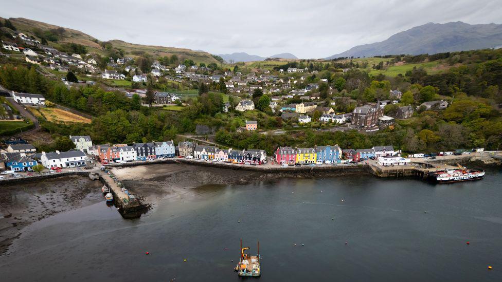 An aerial view of the Isle of Mull, including the port, with brightly coloured buildings at the sea front and sprawling hills in the distance