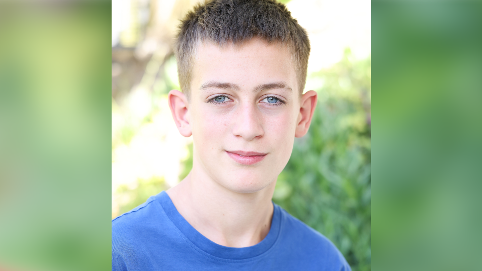 A blonde boy with blue eyes and wearing a blue T-shirt smiling at the camera with vegetation in soft focus behind him.