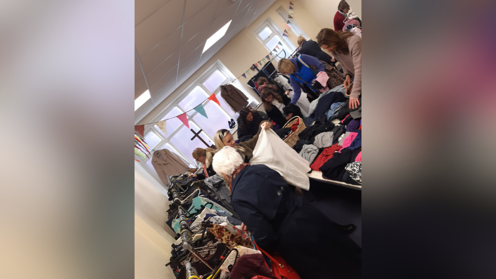 People rummage through a small room containing rails and tables full of clothes. 