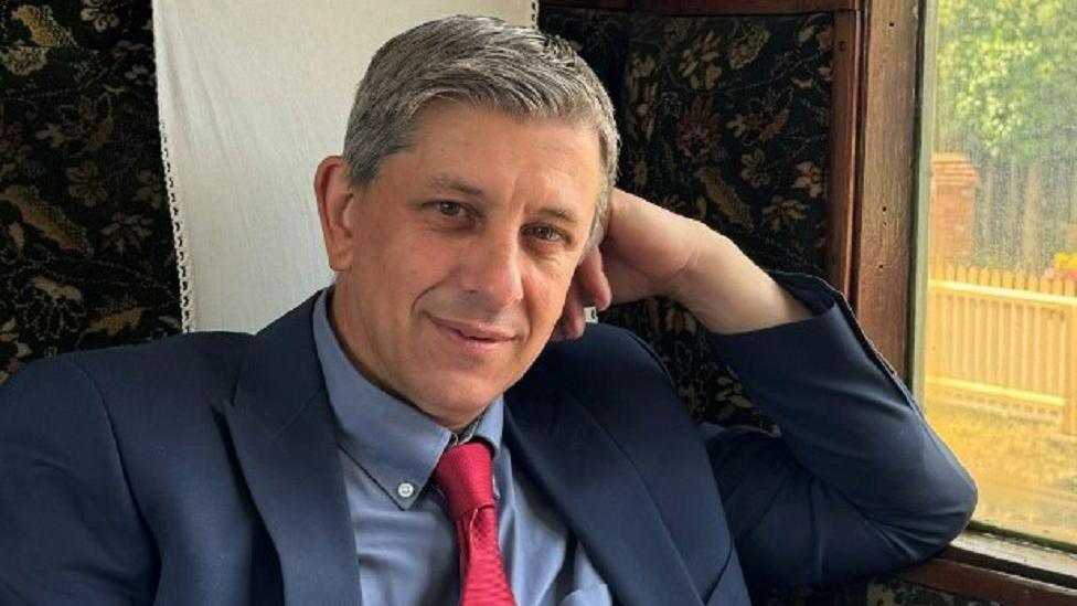 Marc Morgan Huws, chief executive of the Isle of Wight Steam Railway, sitting in a train carriage with his head resting on his left arm and smiling at the camera. He is wearing a navy suit with a light blue shirt and a red tie. 