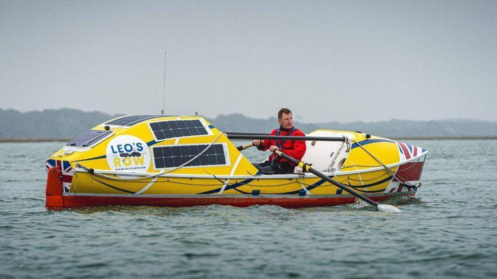 Man in red coat in yellow and red rowing boat he is holding the oars and looking to his left as he moves through the water.