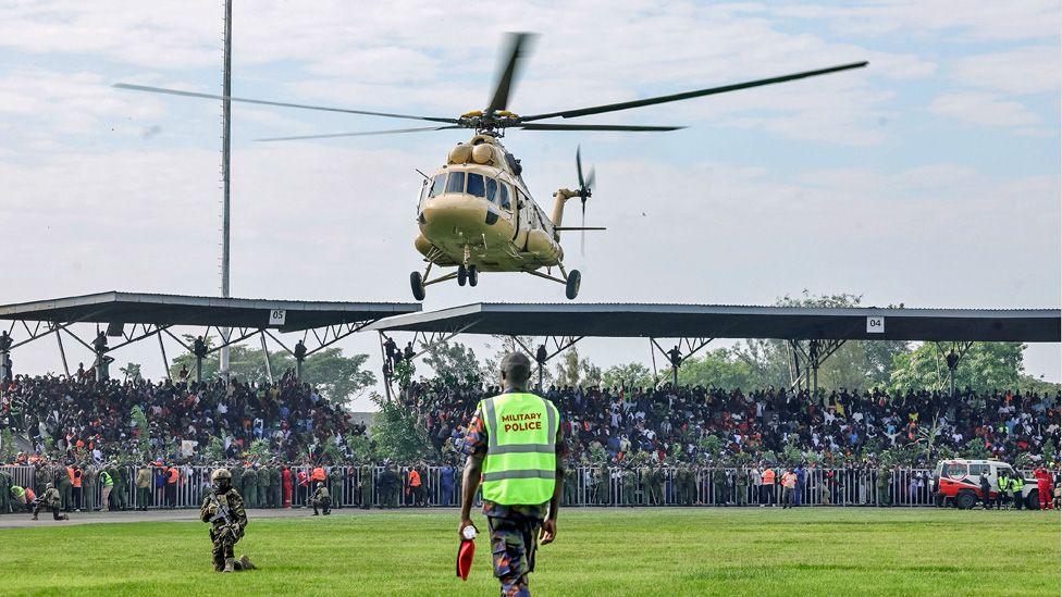 A helicopter about to land on green grass at a stadium in Kisumu. Crowds can be seen behind and security officers on the grass.