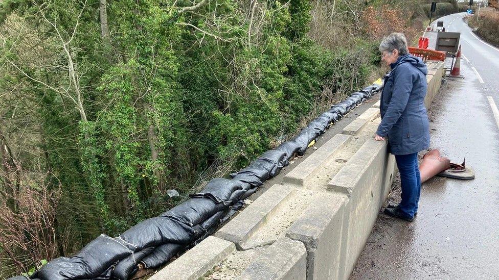 Heather Kidd is wearing a navy blue coat and is standing against concrete bollards that block off a landslip. There are dozens of black sandbags on the edge of a road, with a drop that reveals thick trees. In the background are red roadworks and cones.