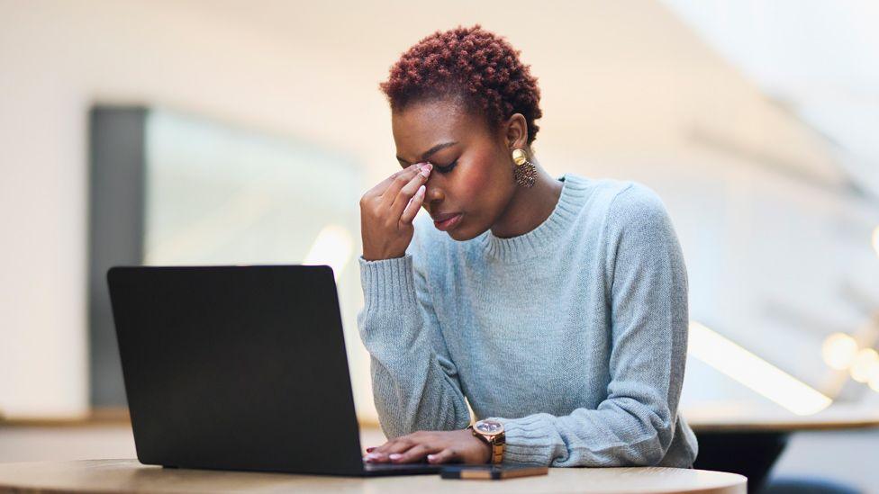 Stock image of a woman sitting at a laptop looking down with her head in her hand. She has short hair and is wearing a blue-grey jumper.