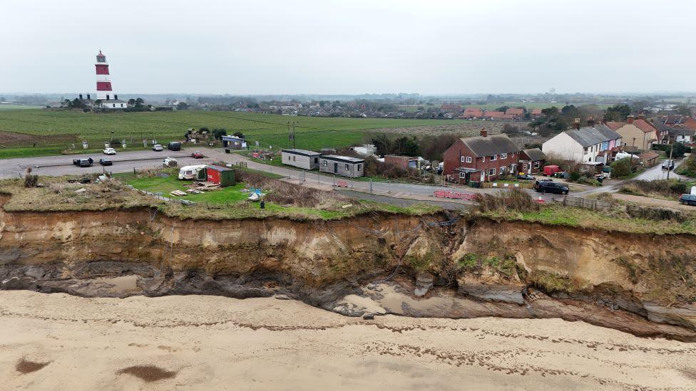 Picture shows the cliff edge at Happisburgh. The cliffs are very sandy and there is a beach below. Houses and cars are very close to the cliff edge and there is a  red and white striped lighthouse in the distance.