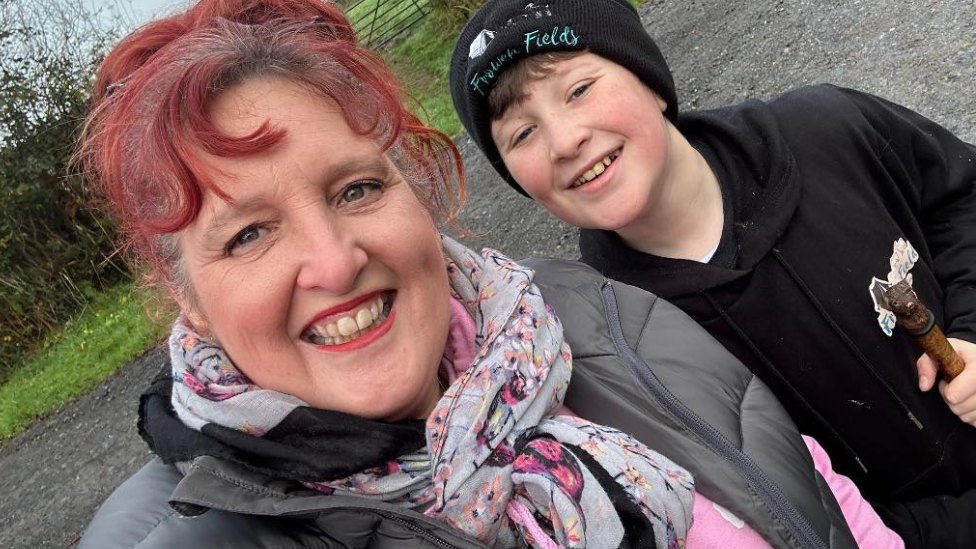 Woman with died red hair in a bun, wearing a grey puffy jacket and gre and pink scarf. Next to her is a 12-year-old boy with brown hair wearing a black hoodie and black beanie. They are both looking at the camera and smiling.