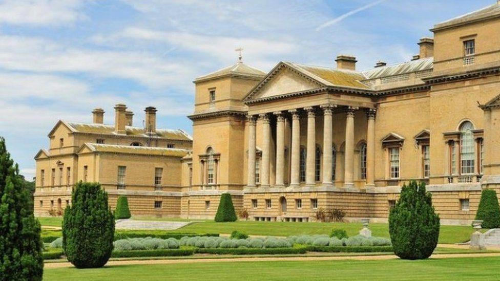 Holkham Hall, an 18th Century mansion. It is built in yellow stone and in front of it are formal gardens of green grass with low box hedges surrounding pale green shrubs. There are also clipped evergreen shrubs.