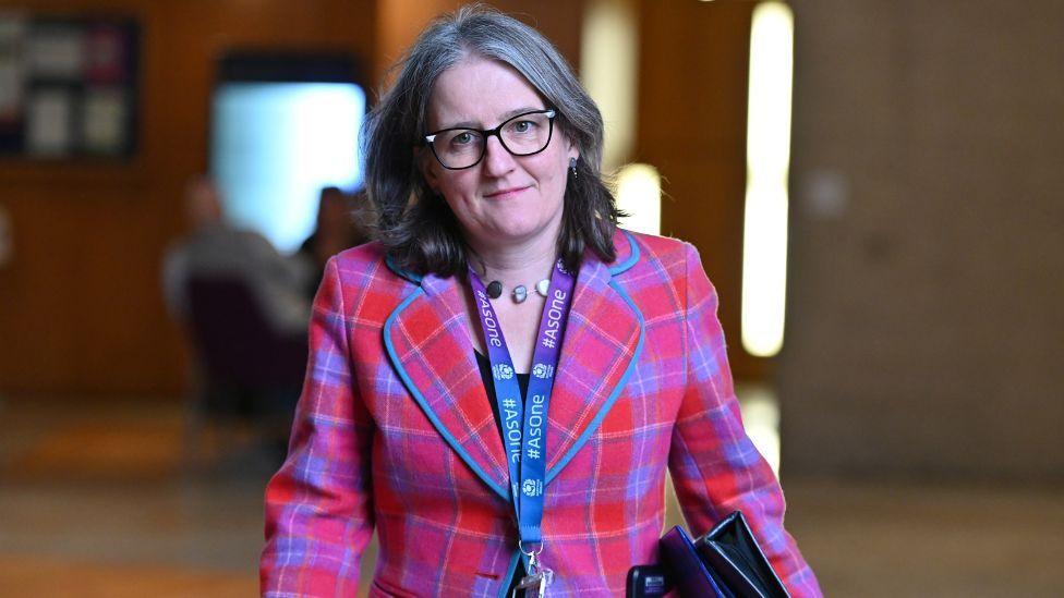 Maree Todd wearing glasses and a pink and blue tartan blazer walking through the Scottish Parliament