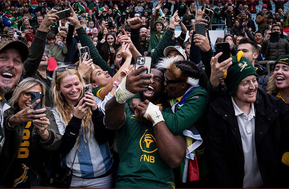 South Africa's Siya Kolisi poses for a selfie with members of the crowd as he celebrates following the team's victory during The Rugby Championship match against Argentina at the Allianz Stadium in London. Kolisi, in a green rugby jersey, holds a smartphone for a selfie among cheering fans. Many raise phones and flags, with stadium seating and banners visible in the background.