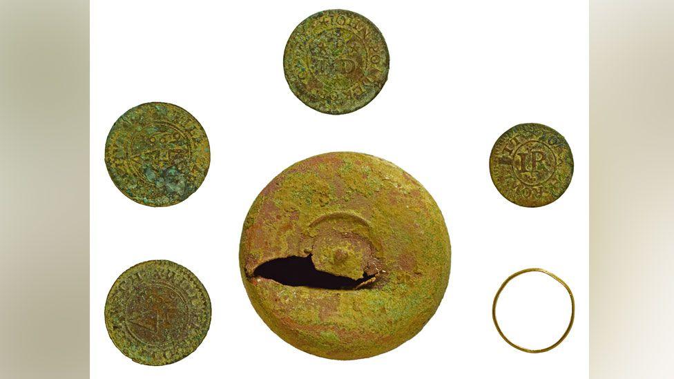 A copper alloy token box with four copper alloy trading tokens and a gold ring. The trading box, which has a gouge out of it, is in the middle and the tokens and ring are placed around the box, evenly spaced.