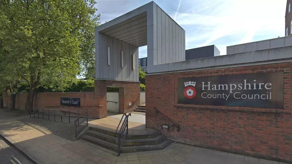 Google street view image of hampshire cc's office. It has two red brick walls with the councils name on signage. There is a gap between the walls which is covered by a silver structure.