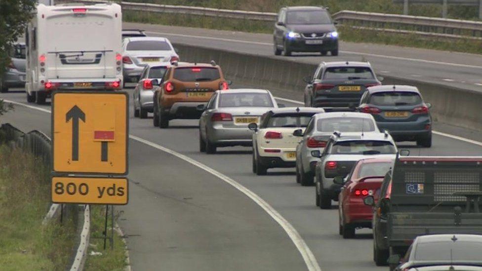 Cars queuing on the M11 motorway. There is a yellow sign to the left which shows there is a lane closure.