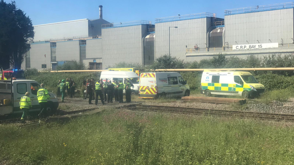 A rail track with many police officers and emergency services vehicles on it. Behind the track are industrial buildings and in front is a grassy verge.