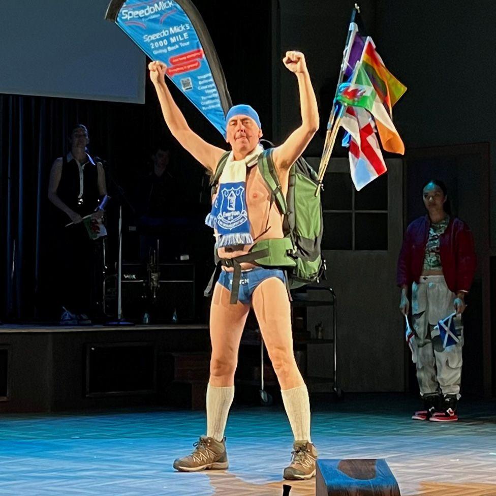 Paul Duckworth as Speedo Mick on stage in a pair of blue SPeedos and Everton scarf with his arms in the air