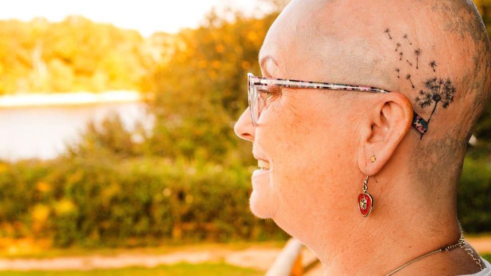 Mandy Nicolson looking out towards a lake at sunset. She has a shaved head and a dandelion tattoo above her left ear