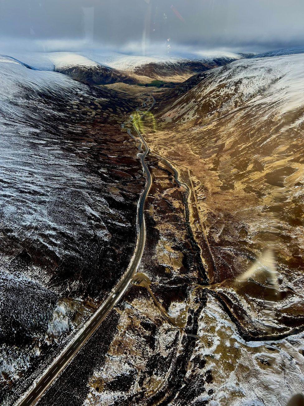 An aerial view of a road cutting through a valley between two snowy hillsides.