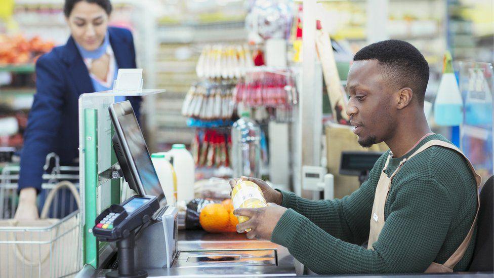 A young man wearing a green sweater and a beige apron works on a supermarket checkout. He is serving a woman wearing a blue and pink top and navy jacket.