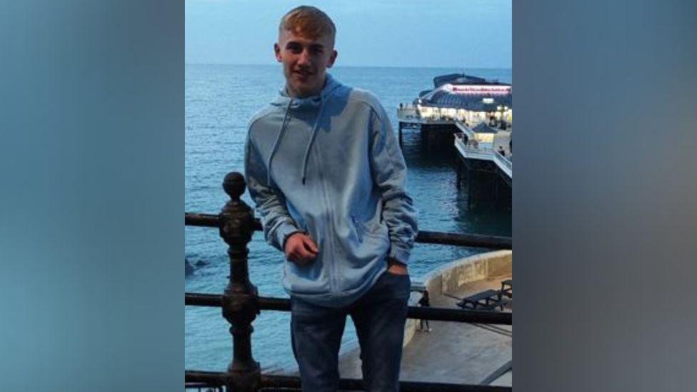 A man wearing a blue zip up hoody is stood with an elbow leaning on a railing with the sea and a pier behind him.