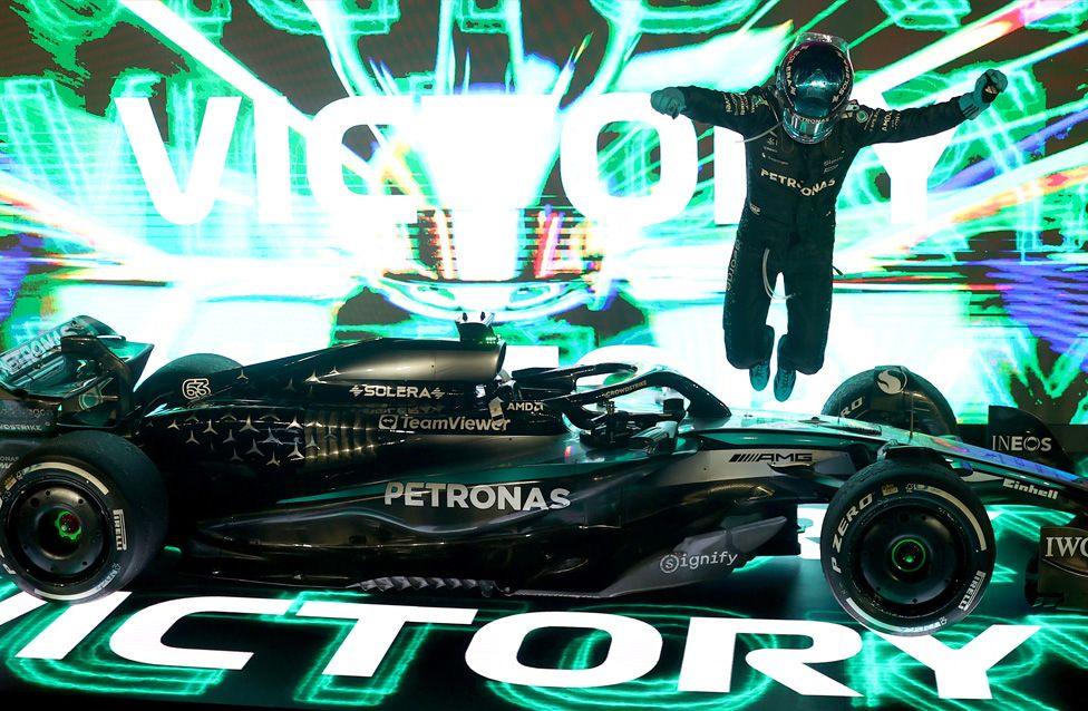 George Russell leaps from his Mercedes after winning the Grand Prix of Singapore. Russell, in a black Petronas racing suit and helmet, jumps beside a sleek black Mercedes Formula 1 car. Behind them, a glowing backdrop displays “VICTORY” in bold letters with bright green and white light effects.