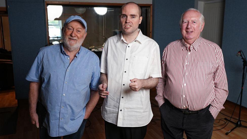 Tony Allen, wearing a blue shirt and blue cap with a white beard, and Mick Fosterm, wearing a red and white striped shirt, stand either side of Martin Finn, who wears a white short-sleeved shirt