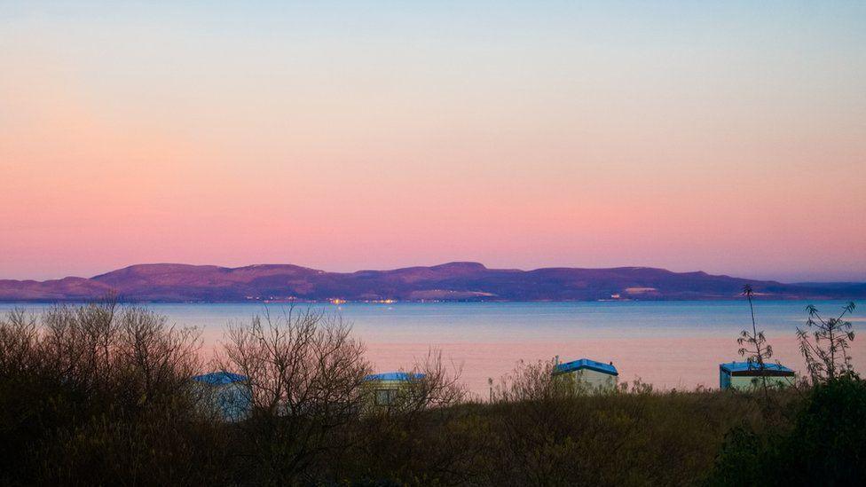 The sky is a firey red colour. There are a few dark hills in the background and four rowing boats in the foreground which are blue and white.