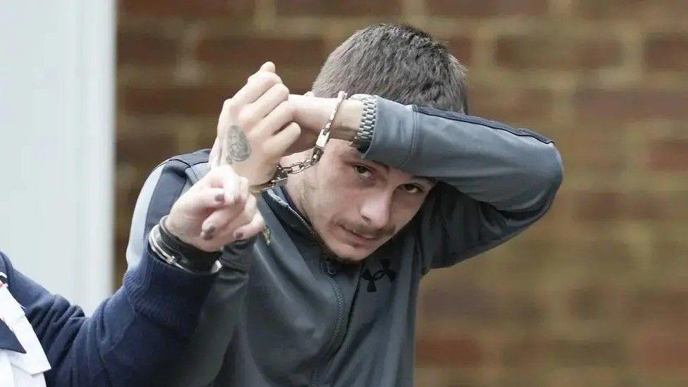 Kyle Dumble in handcuffs. He is attempting to cover his face. He is wearing a grey tracksuit top. 