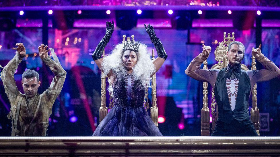 Three professional dancers on Strictly Come Dancing, dressed as skeletons for Halloween week, sitting at a desk with their hands on the table
