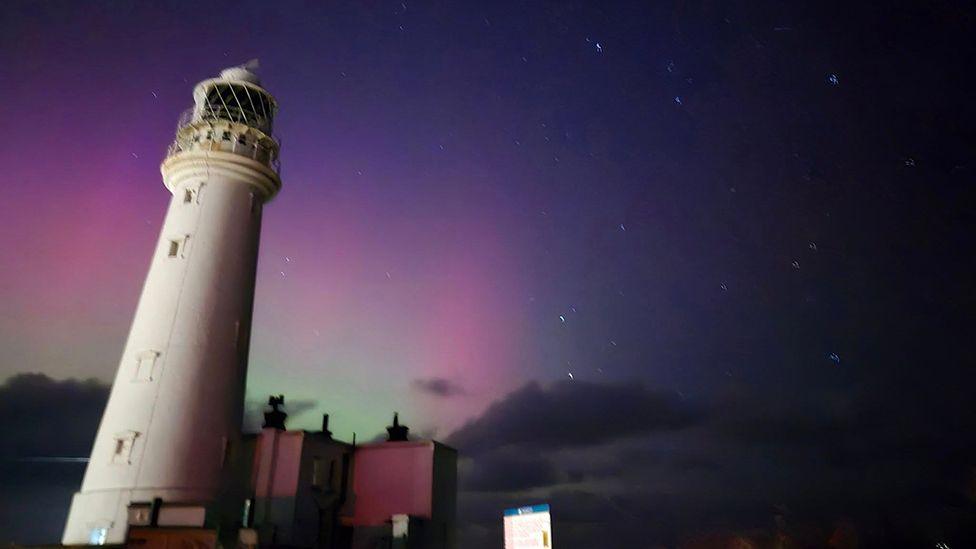 The white lighthouse in Flamborough stands in the foreground with the lights visible above clouds in the distance