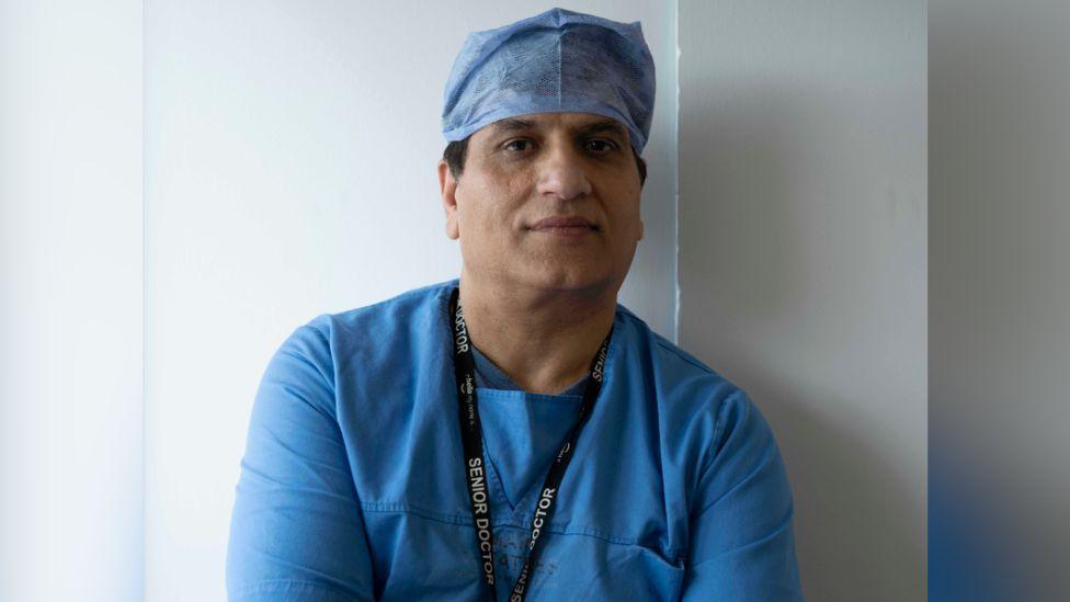 A man in blue medical scrubs looks at the camera, around his neck is a lanyard which says 'senior doctor'.