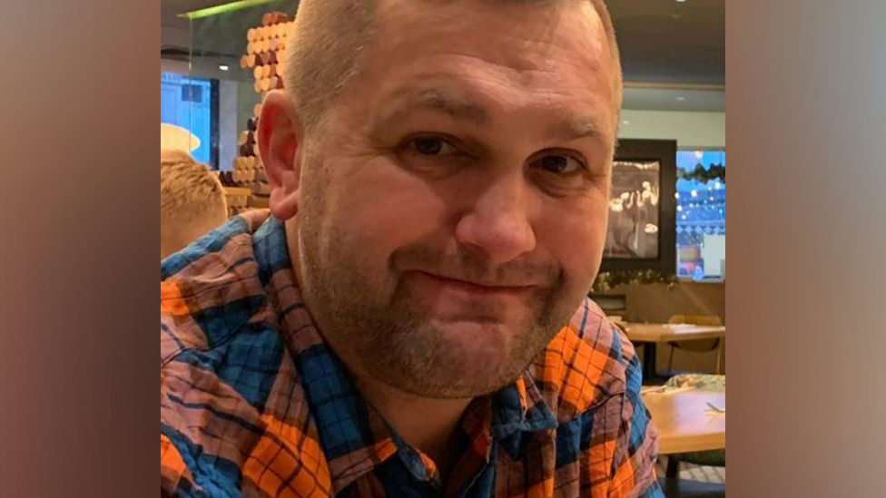 Tomasz Samel pictured wearing an orange and blue a checked shirt. He is smiling and looks to be sat in a pub. He has short dark blond hair and a stubbly beard.