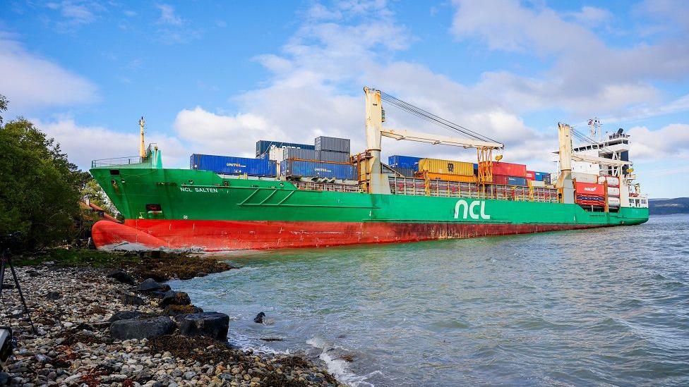 A side view of the ship, taken from shore, shows the front of the boat run aground on a pebbly beach