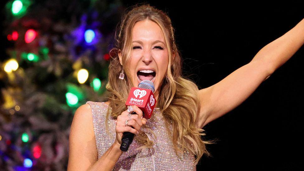 Nikki Glaser apparently shouting into a microphone with her arm raised, at the iHeartRadio Jingle Ball in Inglewood, California, on 6 December