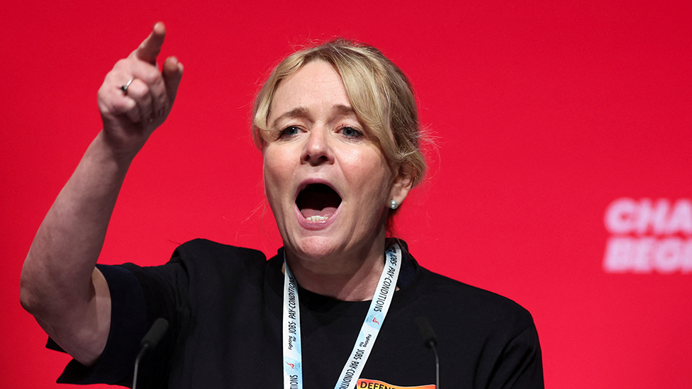 A woman with blonde hair tied back in a ponytail stands in front of a red background. She is wearing a black top and has a lanyard round her neck. She is gesticulating with her hand raised and forefinger pointing and she appears to be shouting or at least talking animatedly.