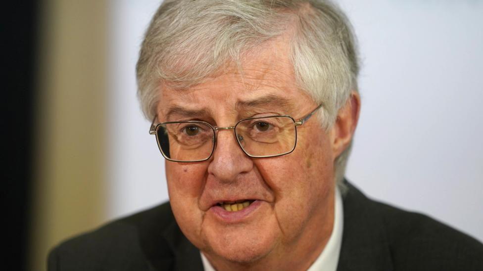 A head and shoulders shot of Mark Drakeford wearing a black suit and white shirt in front of a white background.