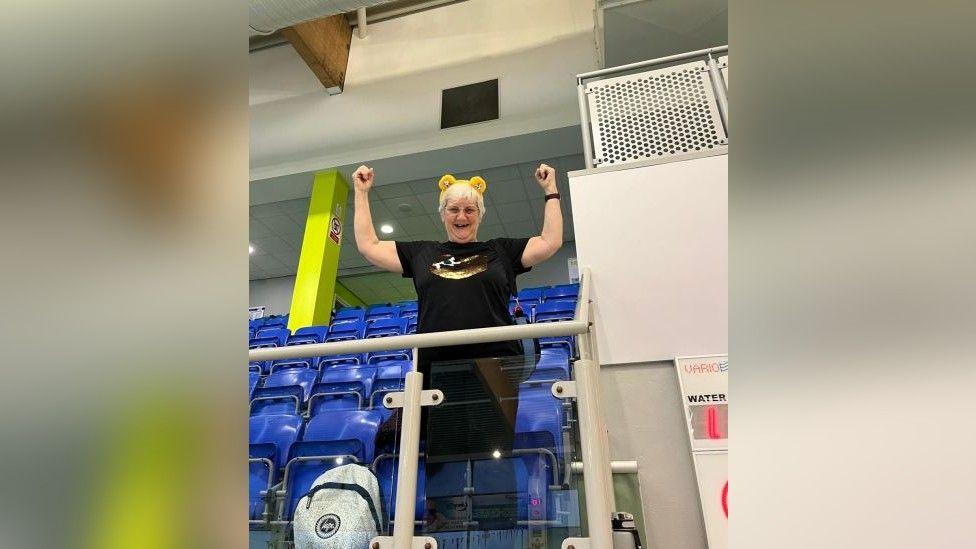 A woman holding both fists in the air and smiling while standing next to several chairs in a viewing area that look out at a swimming pool. She is wearing glasses, has short white hair and is also wearing yellow Pudsey ears.