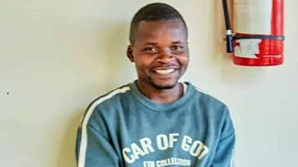 A head and shoulder image of Shedrack Chaula smiling at the camera. he is wearing a green sweatshirt and part of a fire extinguisher can be seen on the wall behind him.