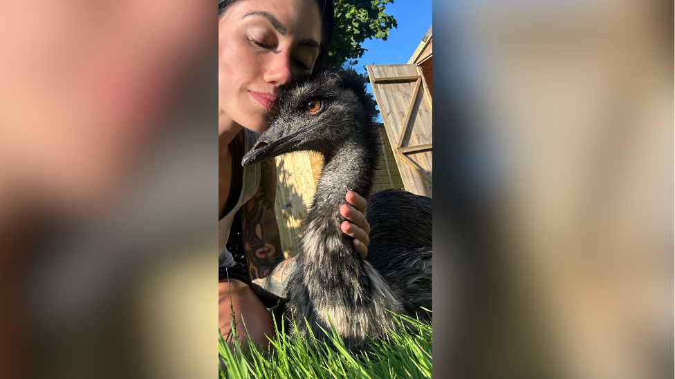 Rhi Evans lies on the grass with emu Jonathan. She has her cheek against the bird's head.